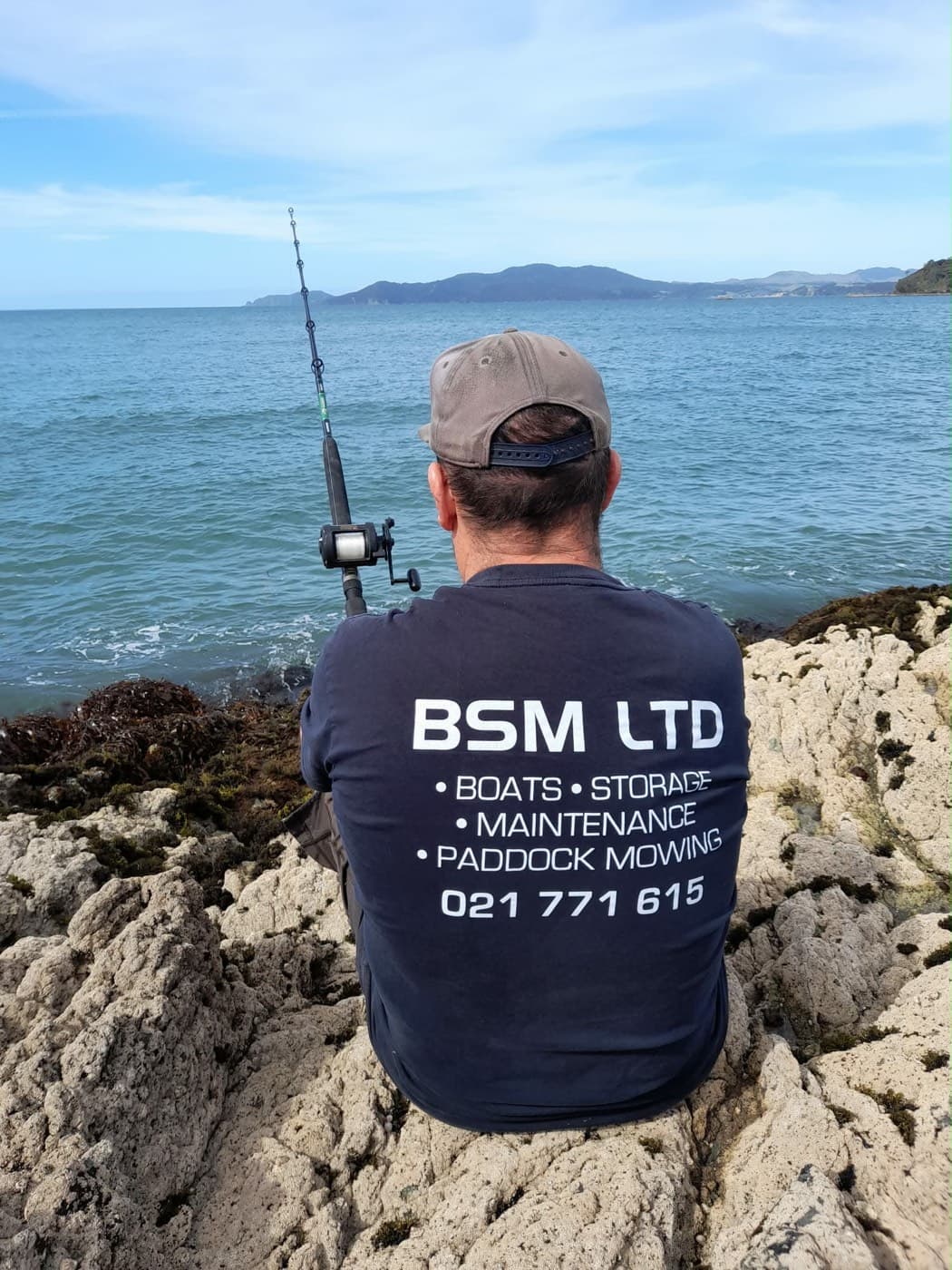 BSM Ltd shirt on the rocks with a fishing rod, looking out over the coast